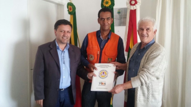 três homens de pé seguram uma pasta com o logo da Defesa Civil
