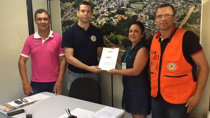 No centro da foto um homem recebe das mãos de uma mulher alguns papéis. Ao lado de ambos posam dois homens. Estão em uma sala com o mapa da cidade ao fundo