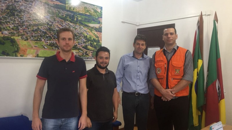 Quatro homens em pé, com roupas claras. Ao fundo da sala, um quadro com uma foto do município.