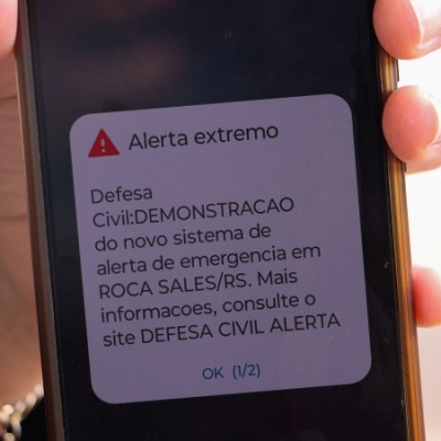Uma pessoa segura um celular, no qual é possível ler Alerta extremo. Defesa Civil: demonstração do novo sistema de emergência em Roca Sales/RS. Mais informações consulte o site Defesa Civil Alerta