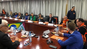 Imagem mostra 13 pessoas participando de uma reunião, as bandeiras do Brasil e do Peru podem ser vistas entre as mesas.