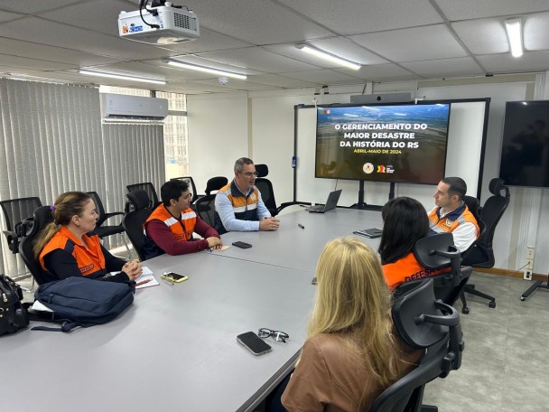 Pessoas da Defesa Civil do Acre e do Rio Grande do Sul estão sentadas ao redor de uma mesa conversando. Ao fundo, tela com o escrito "O gerenciamento do maior desastre da história do RS"