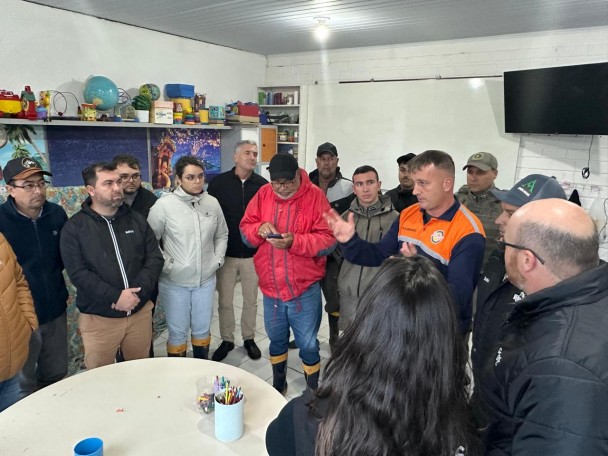 Grupo de 14 pessoas, de pé, em uma sala de aula, ouvindo um homem, vestido com camisa da Defesa Civil Estadual, explicar algo.