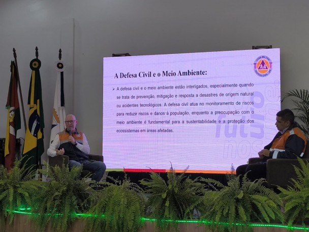 Os representantes da Defesa Civil estão sentados, com plantas em primeiro plano. Ao fundo, tela com slide sobre a Defesa Civil e o Meio Ambiente