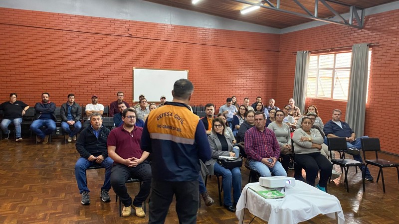 O palestrante, vestindo casaco da Defesa Civil, está de costas, falando para um público sentado