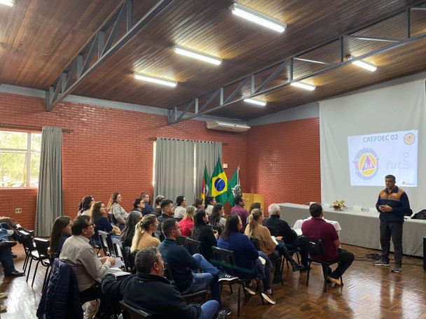 O palestrante, vestindo um casaco da Defesa Civil, está de frente, e podemos ver o público de costas, sentado. Ao fundo, um slide da Crepdec 2 está sendo projetado em uma tela branca.