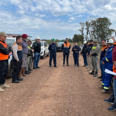 Um grupo de pessoas est&aacute; reunida em roda, em uma estrada de barro. &Eacute; poss&iacute;vel verificar pelas vestimentas que participam de diferentes &oacute;rg&atilde;os, como da Defesa Civil, da Brigada Militar e companhia de g&aacute;s.