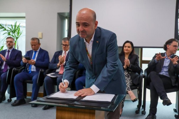 Coronel Boera está assinando o Acordo de Cooperação. Ao fundo, pessoas sentadas aplaudem.